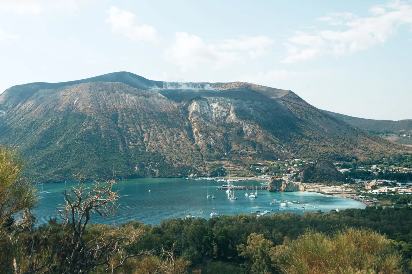 A steaming volcanic crater with a pale sulphur scar down its flank above a turquoise bay of moored yachts