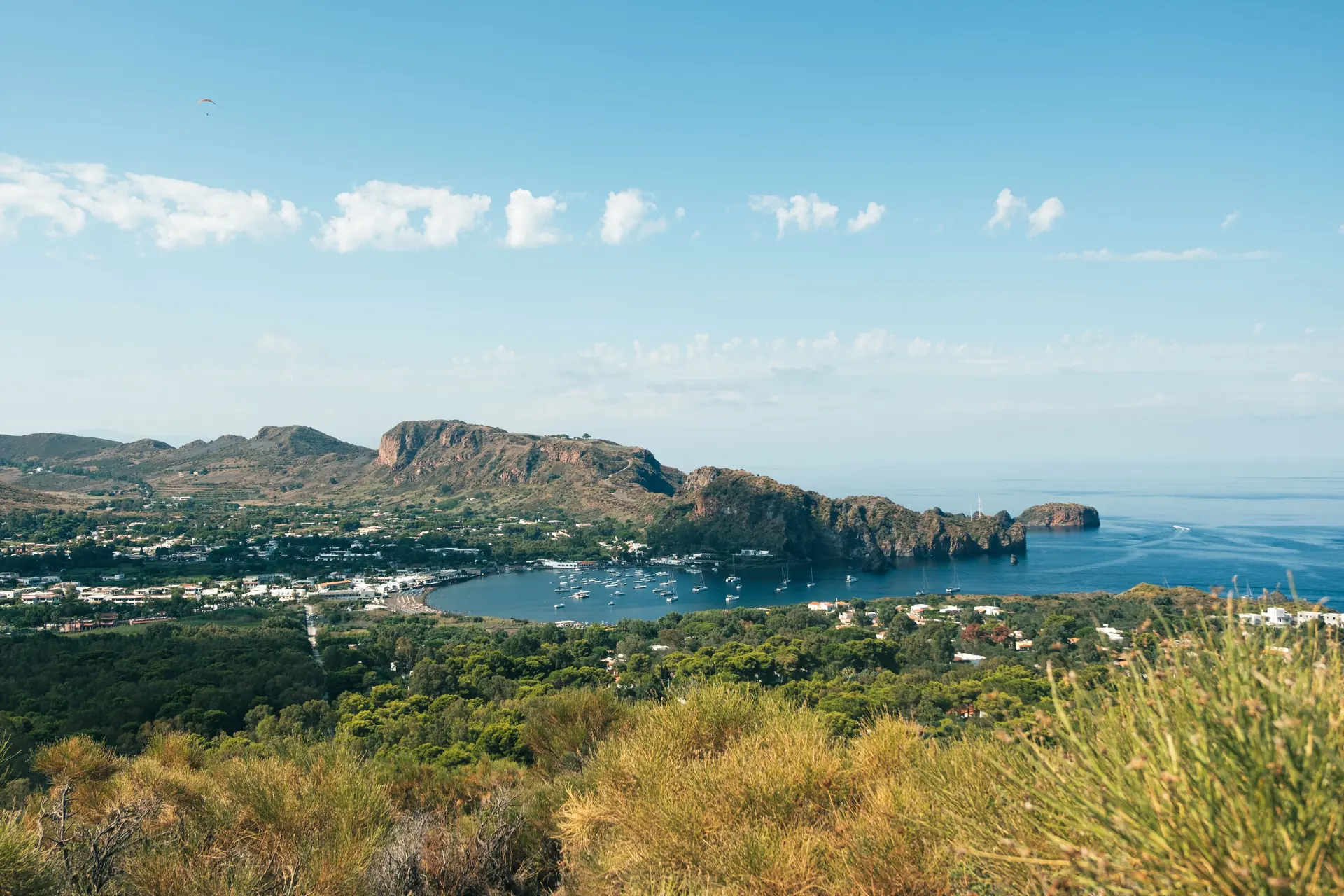 A scrub-covered ridge above a bay of moored sailing boats, white flat-roofed houses and a rust-red lava headland