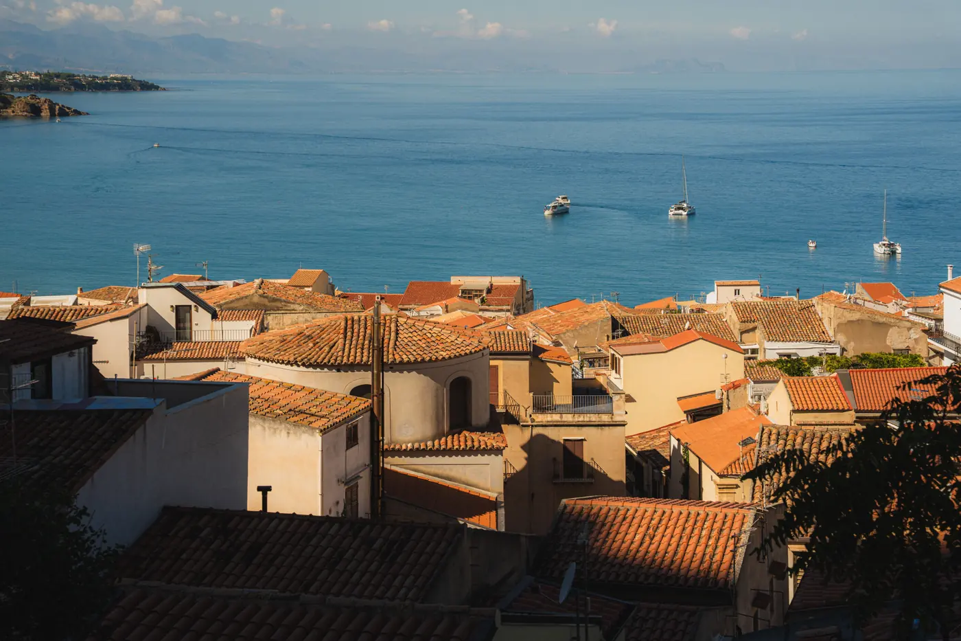 Terracotta-tiled rooftops stepping down to a blue bay with moored sailing boats in low sun