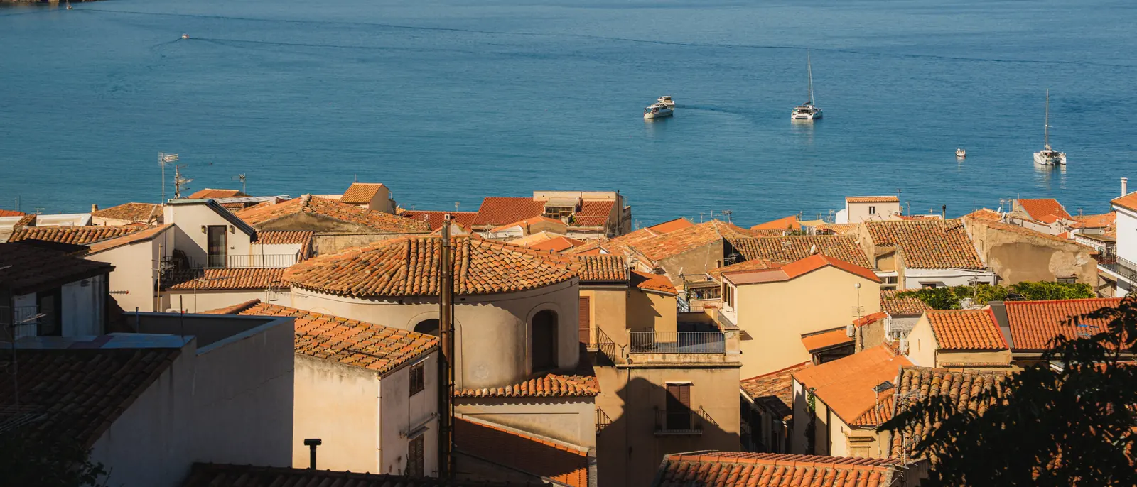 Terracotta-tiled rooftops stepping down to a blue bay with moored sailing boats in low sun