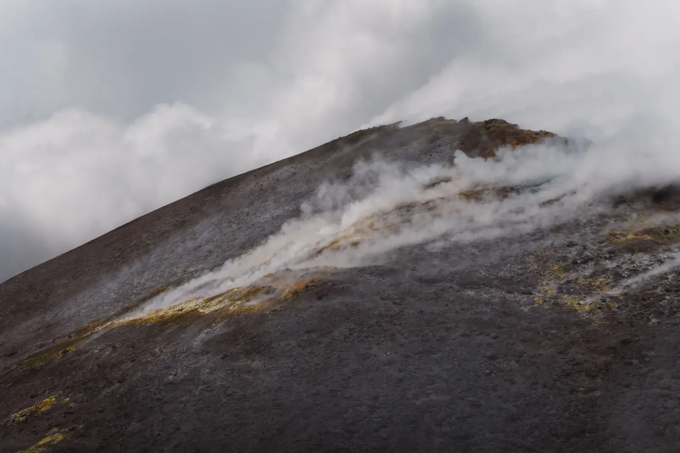 Steam and sulphur venting from the bare grey flank of a volcanic cone under low cloud