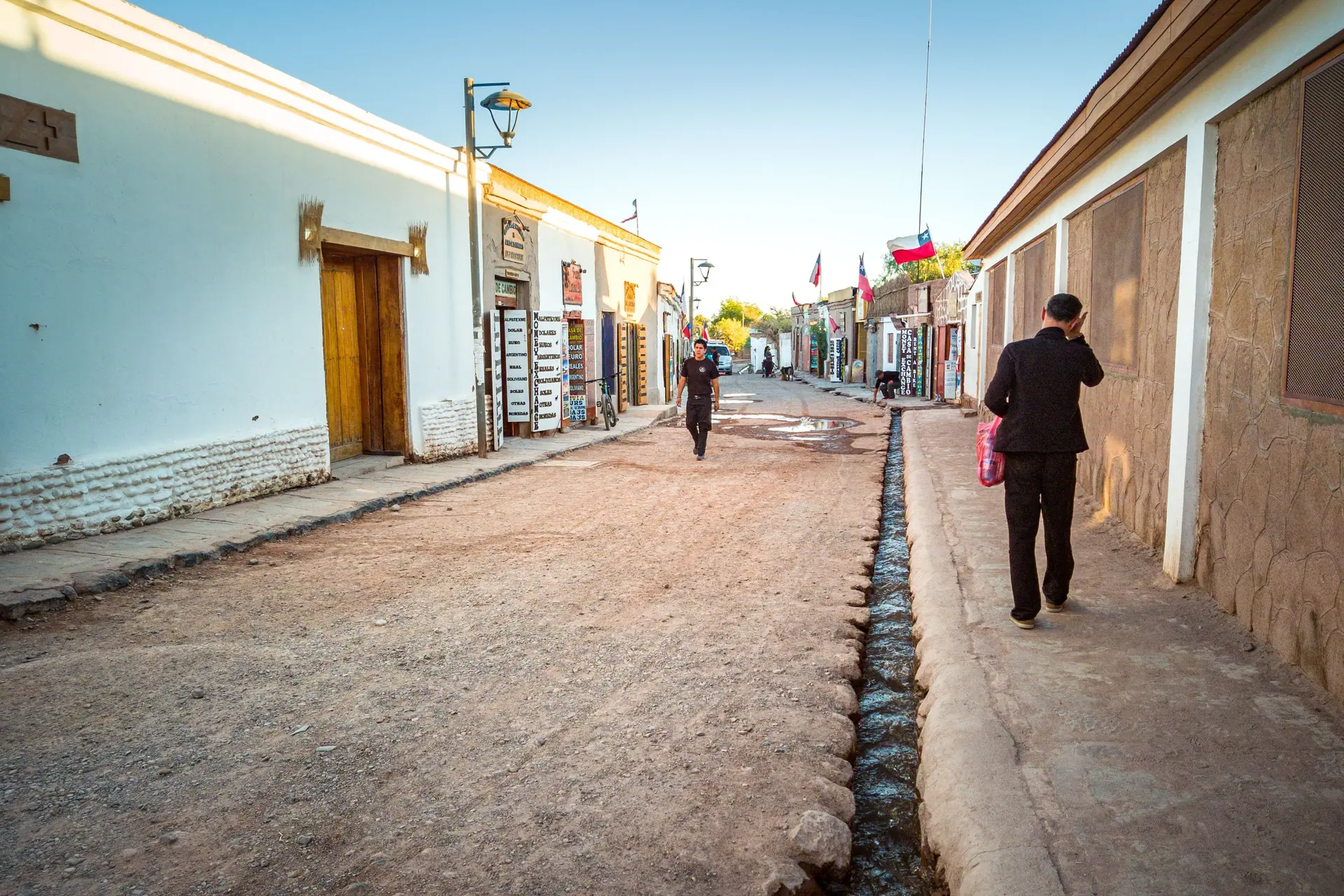 An adobe street in San Pedro de Atacama with two people walking and painted shop boards