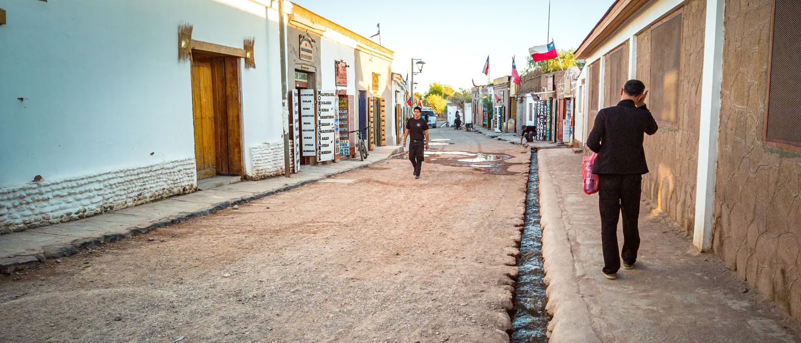 An adobe street in San Pedro de Atacama with two people walking and painted shop boards