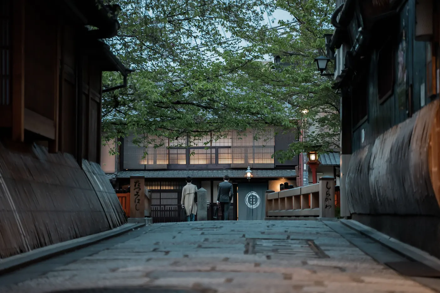 A stone-paved lane in Gion, Kyoto at dusk between wooden machiya with cedar lattice fronts, two people walking away towards a lit bridge
