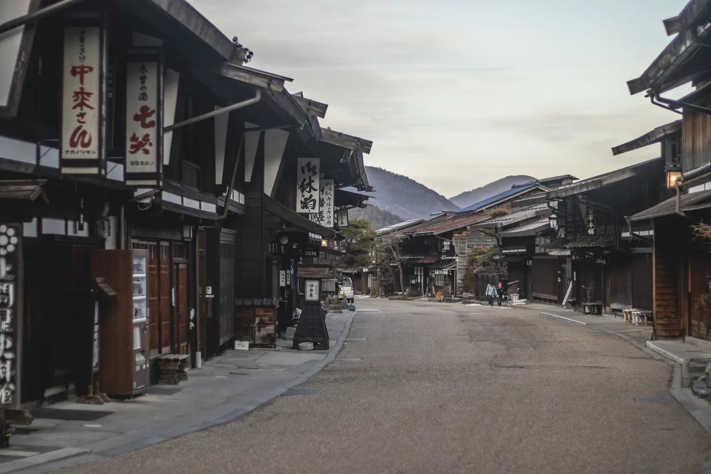 A preserved timber post-town street with deep eaves, two distant figures and a vending machine