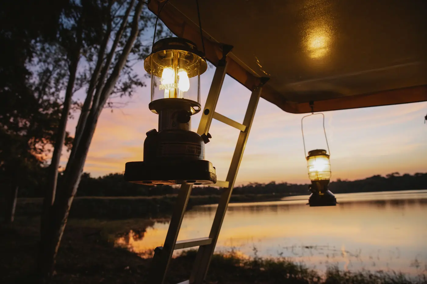 Two paraffin storm lanterns and a step-ladder under a canvas awning above still water at an orange dusk
