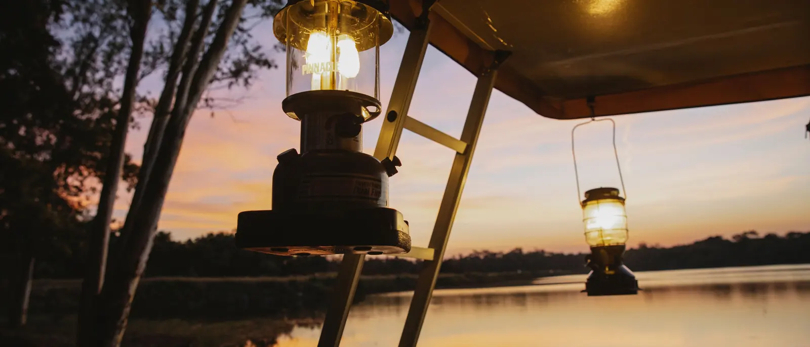 Two paraffin storm lanterns and a step-ladder under a canvas awning above still water at an orange dusk