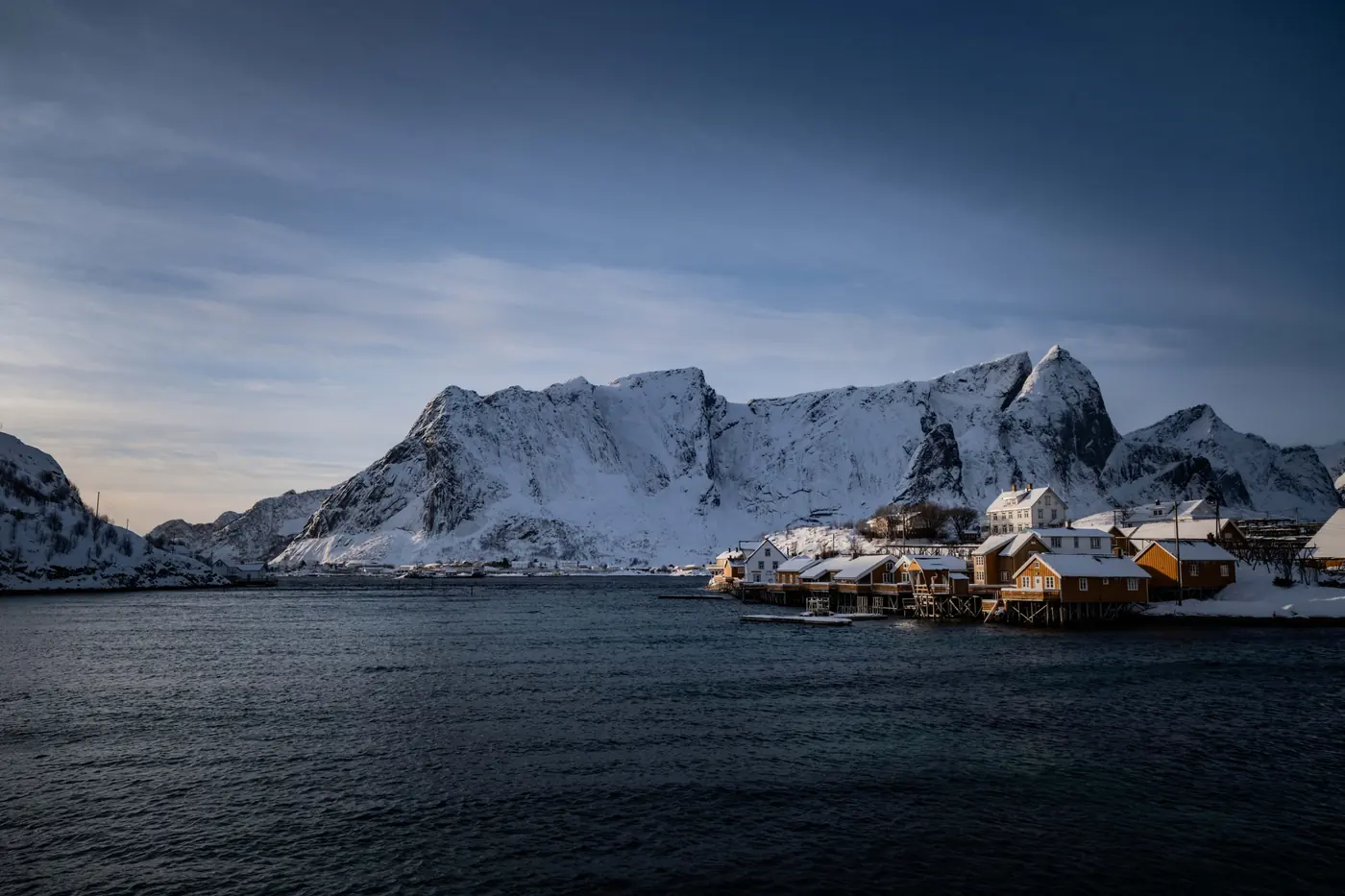 A dark snow-gullied granite peak rising straight from the sea above a row of ochre cabins on stilts