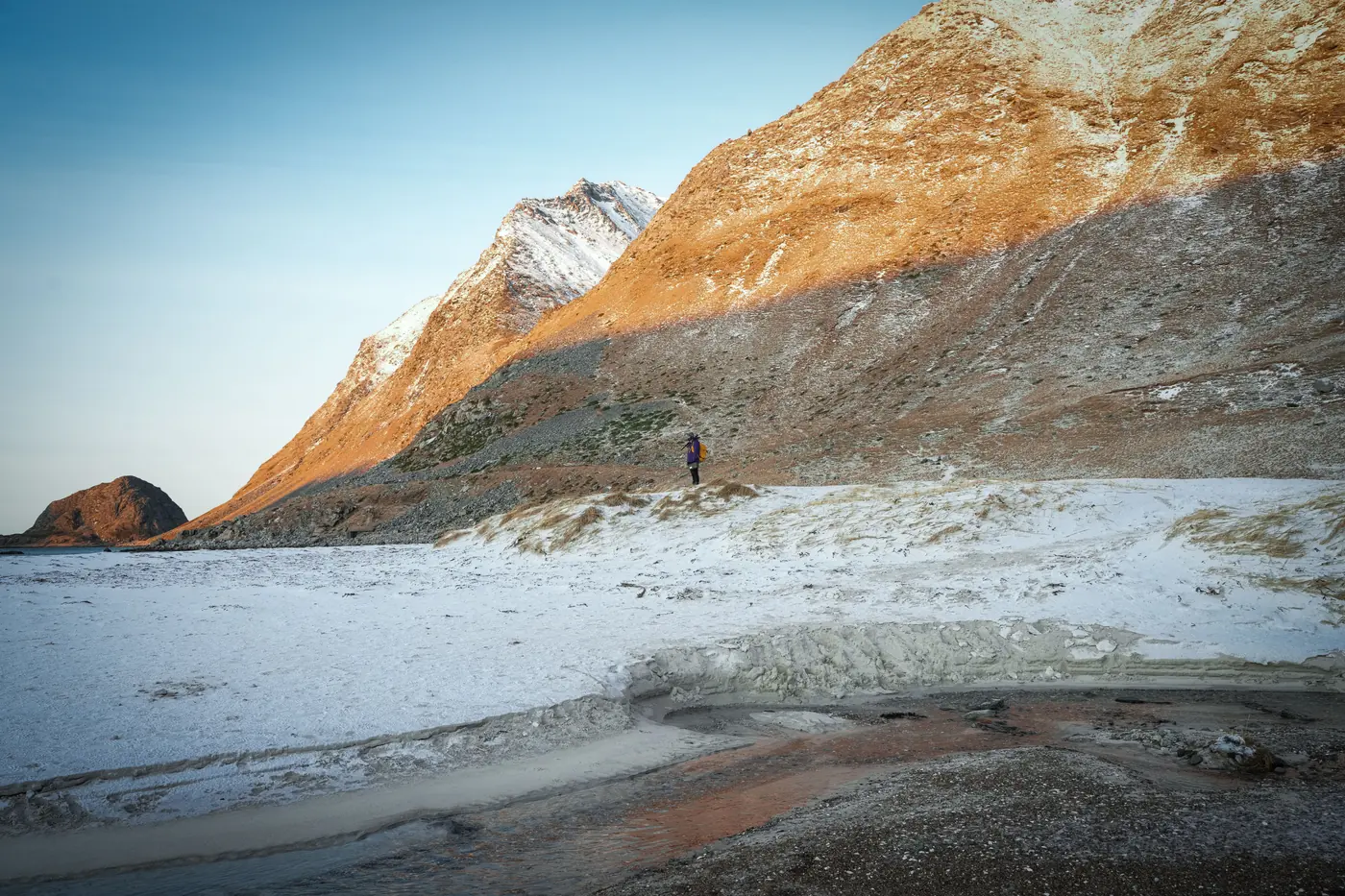Low sun on a bare mountain flank above a snow-covered shore, one distant walker below