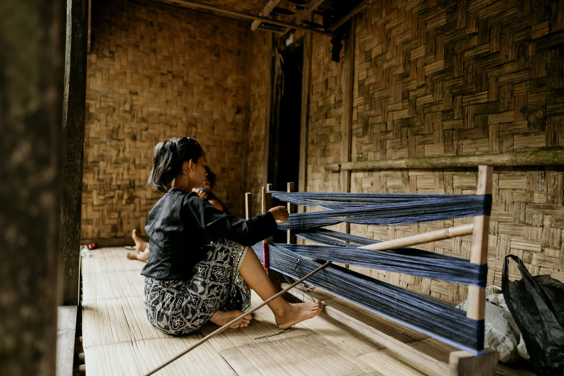 A woman sitting on a split-bamboo floor tensioning an indigo warp on a low backstrap loom