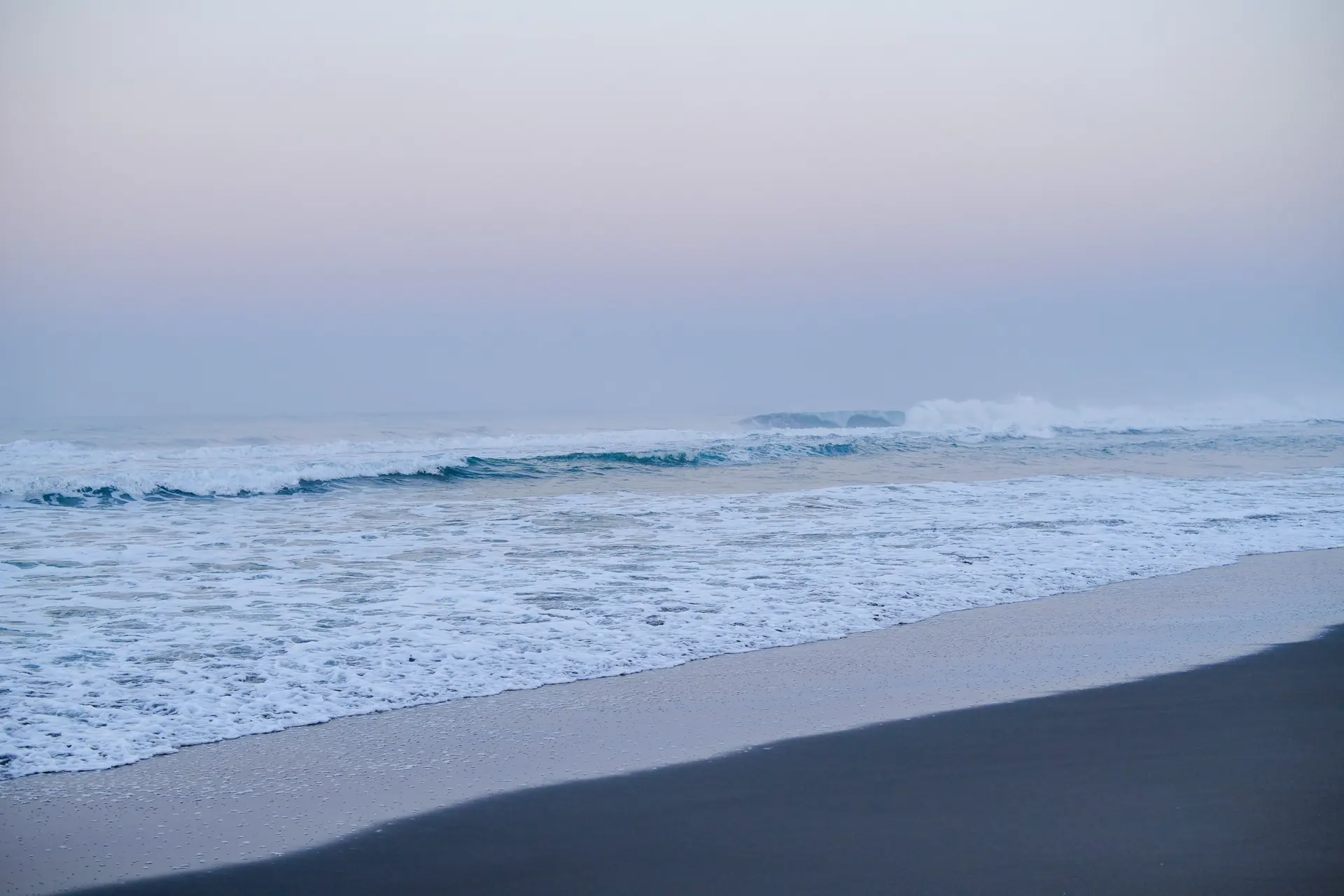 Lines of open-ocean swell breaking onto a completely empty shore under a lavender and pink dawn sky