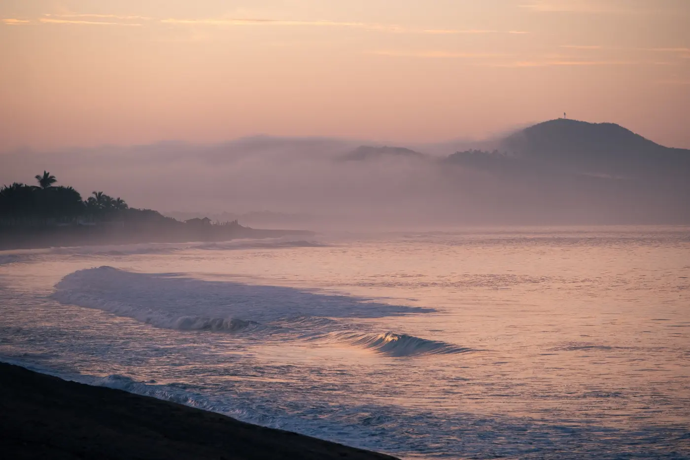 A long line of swell breaking on an empty dark beach below hazy headlands at sunset