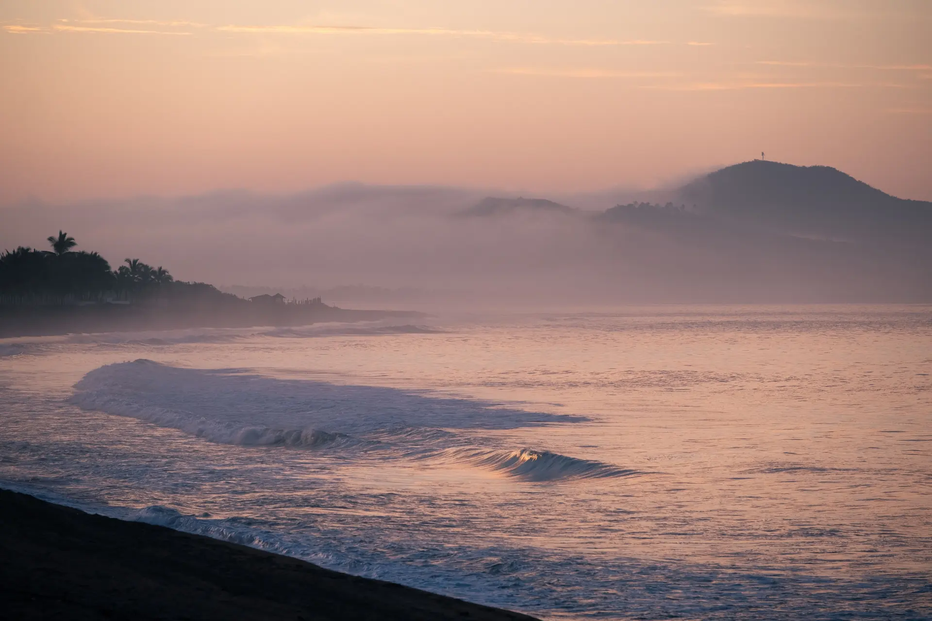 A long line of swell breaking on an empty dark beach below hazy headlands at sunset