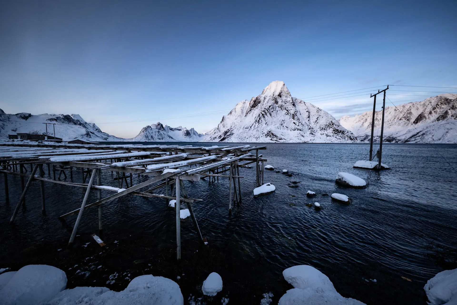 Tall timber cod-drying racks standing in dark water below a snow-covered granite peak at blue hour