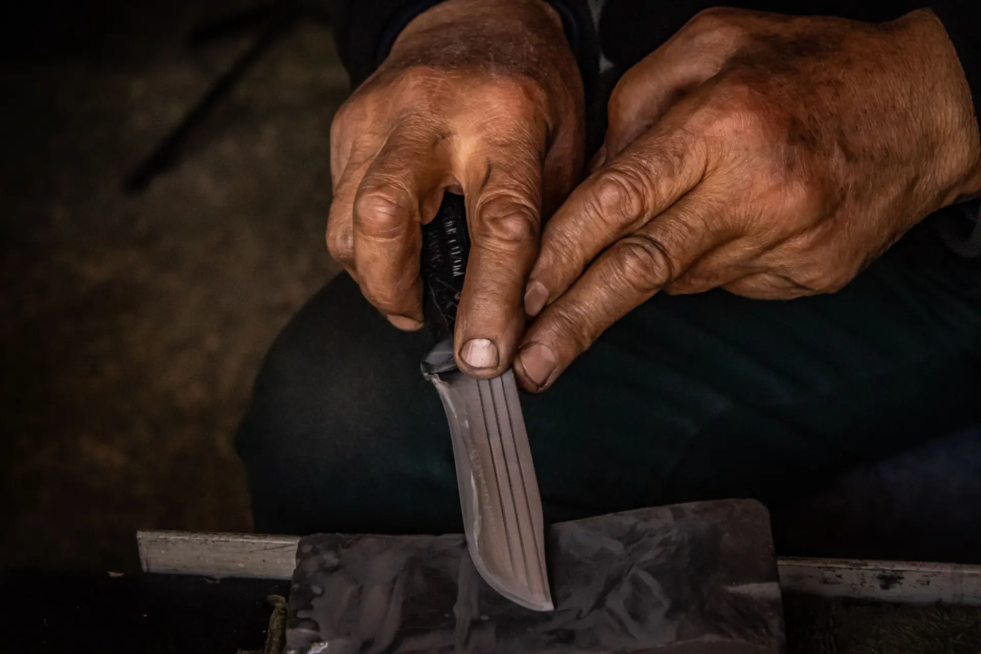 Two weathered hands holding a carbon-steel blade flat against a wet sharpening stone on a timber rest