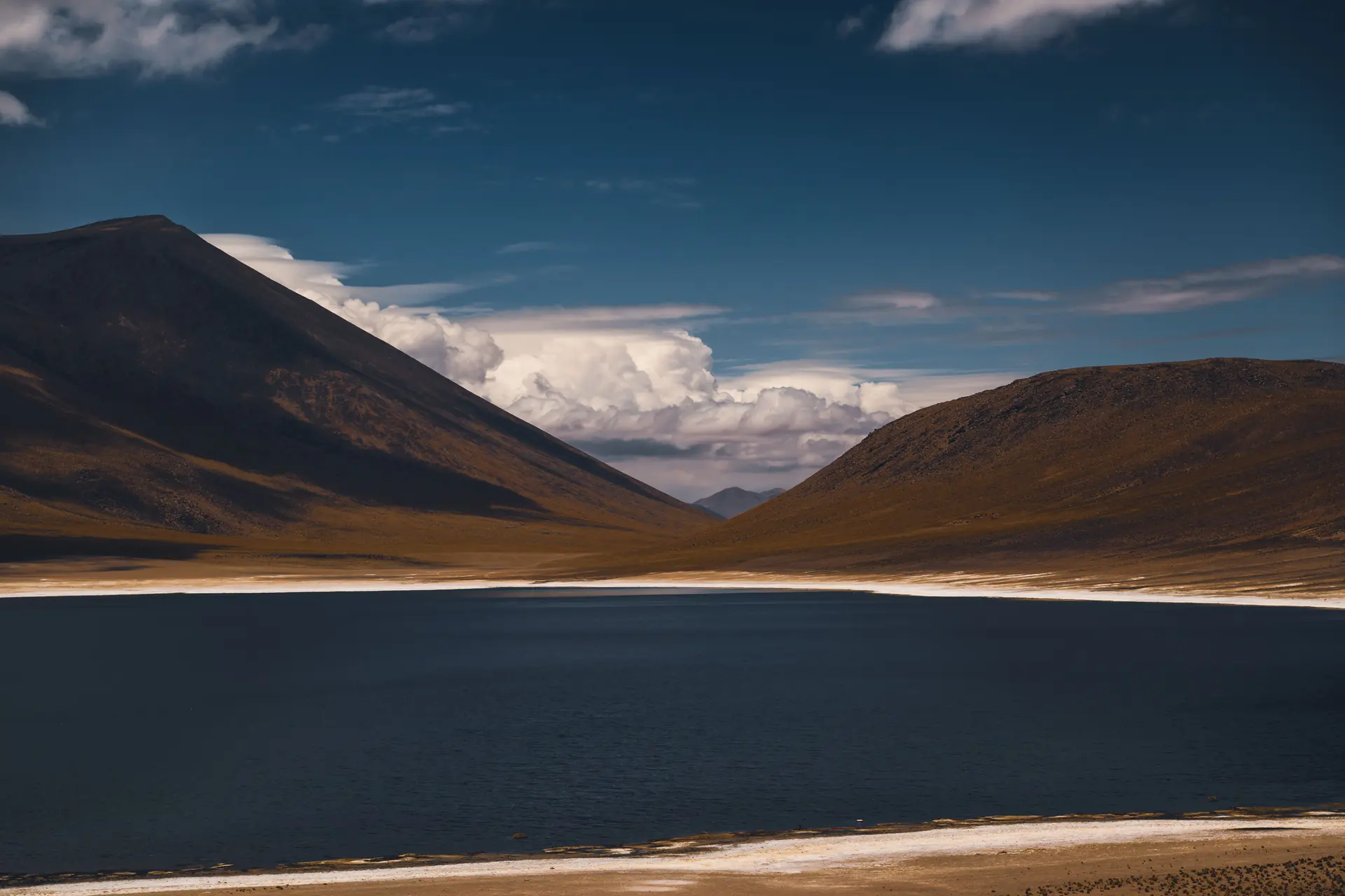 A deep blue altiplano lagoon rimmed with white salt crust between two bare volcanic slopes under afternoon cumulus