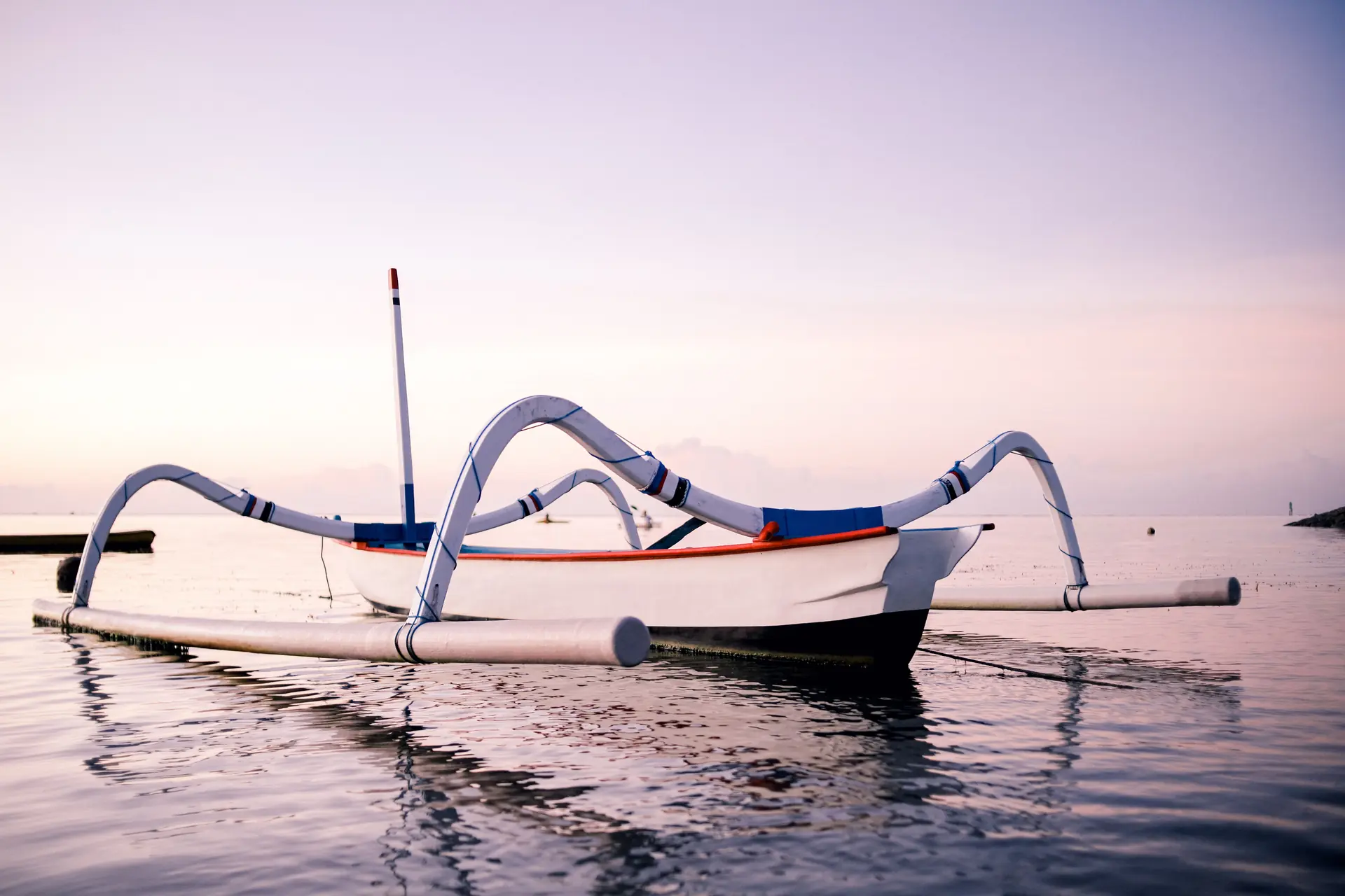 A narrow outrigger canoe with bamboo booms mirrored in glassy water under a lavender dawn sky