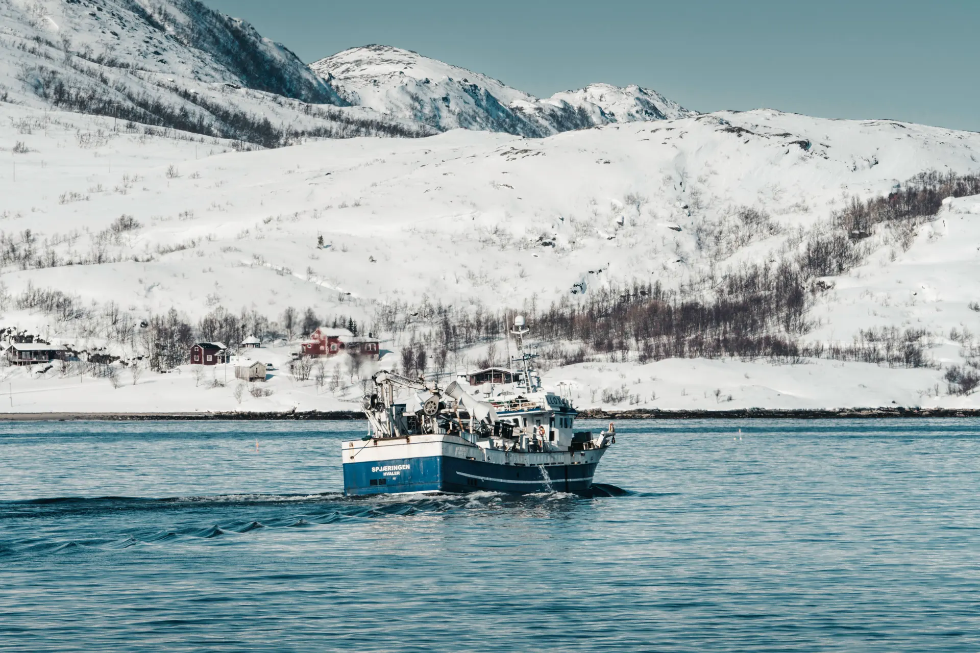 A blue-and-white fishing boat under way toward a snow-covered shore lined with red and white timber houses