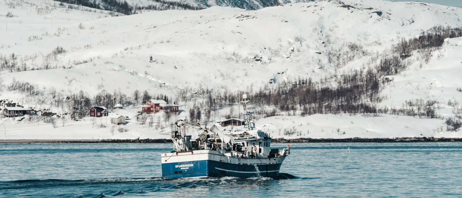 A blue-and-white fishing boat under way toward a snow-covered shore lined with red and white timber houses