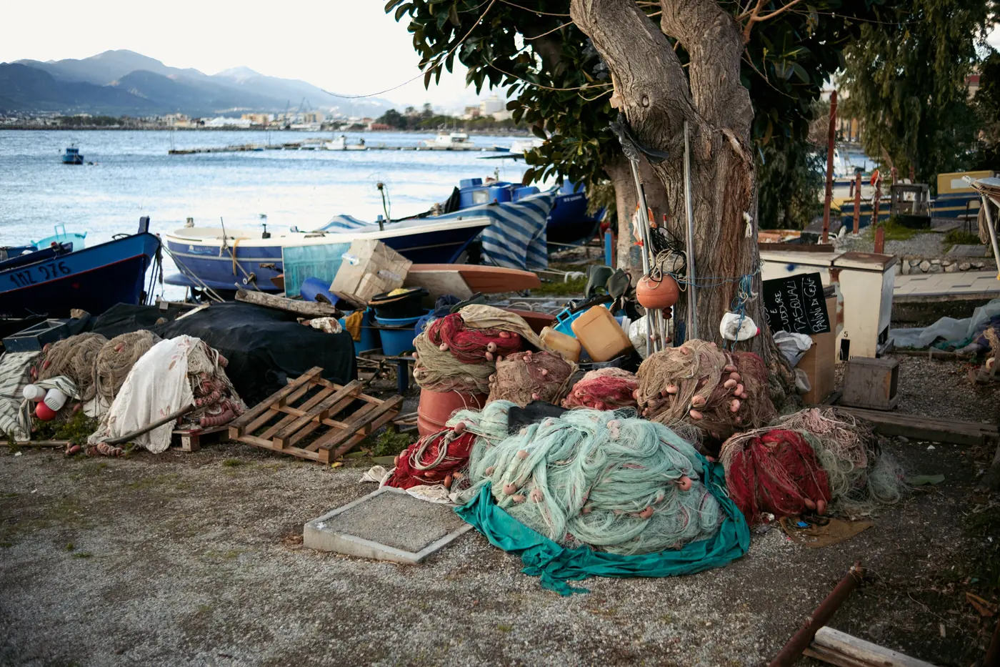 Heaps of jade and rust fishing net piled under a tree on a gravel foreshore, boats hauled up behind