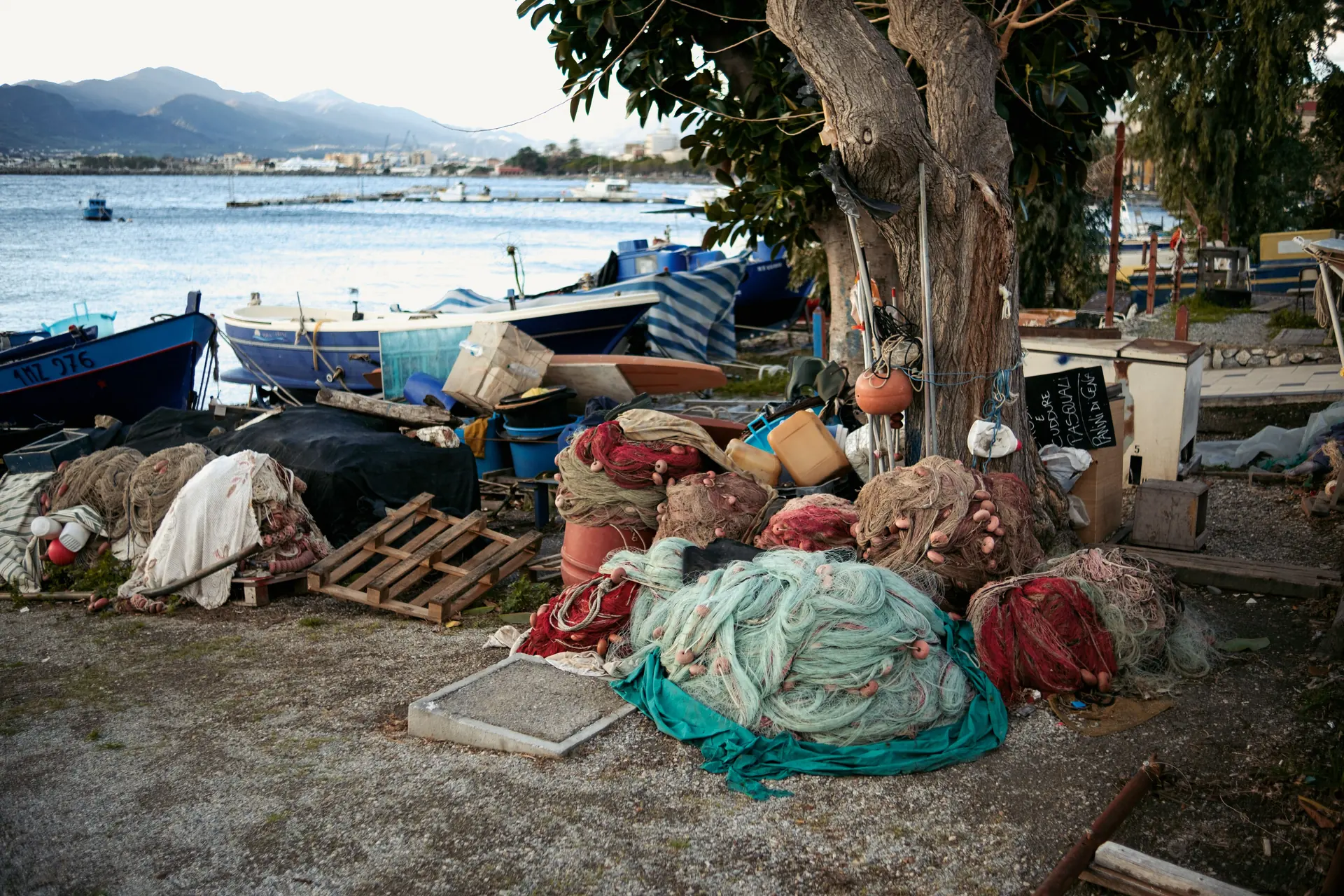 Heaps of jade and rust fishing net piled under a tree on a gravel foreshore, boats hauled up behind
