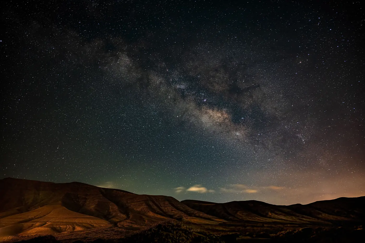 The Milky Way over a bare eroded desert ridge, a dense starfield with faint green airglow near the horizon