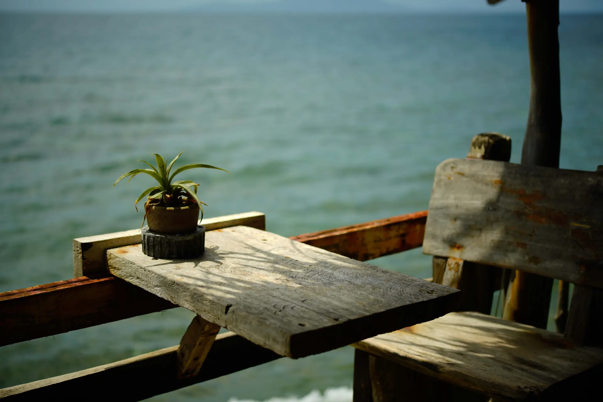 A weathered timber table and plank bench on an open deck above calm teal sea, a headland on the horizon