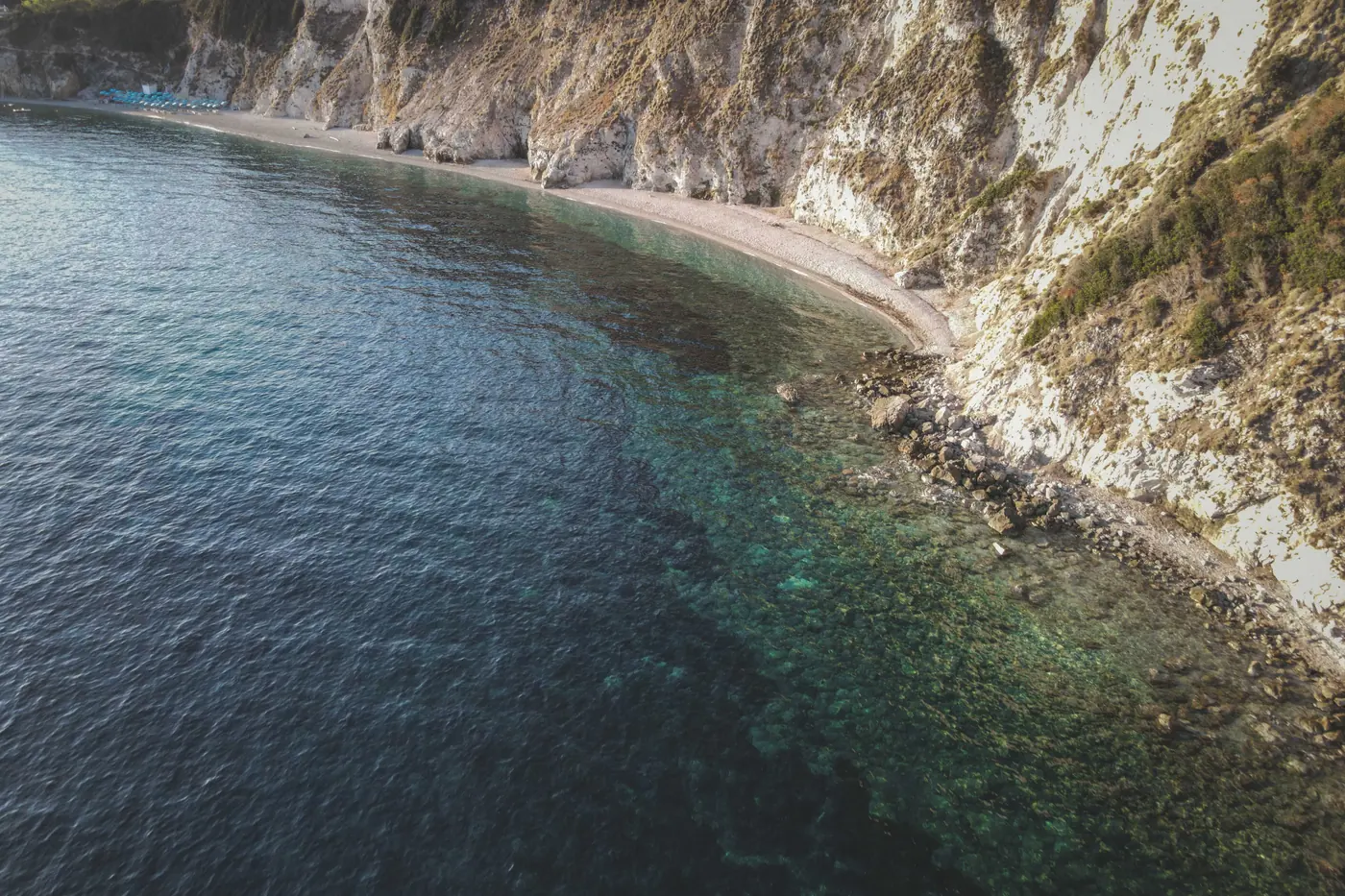 A steep white cliff dropping straight into water that shades from deep indigo to pale jade in low sun