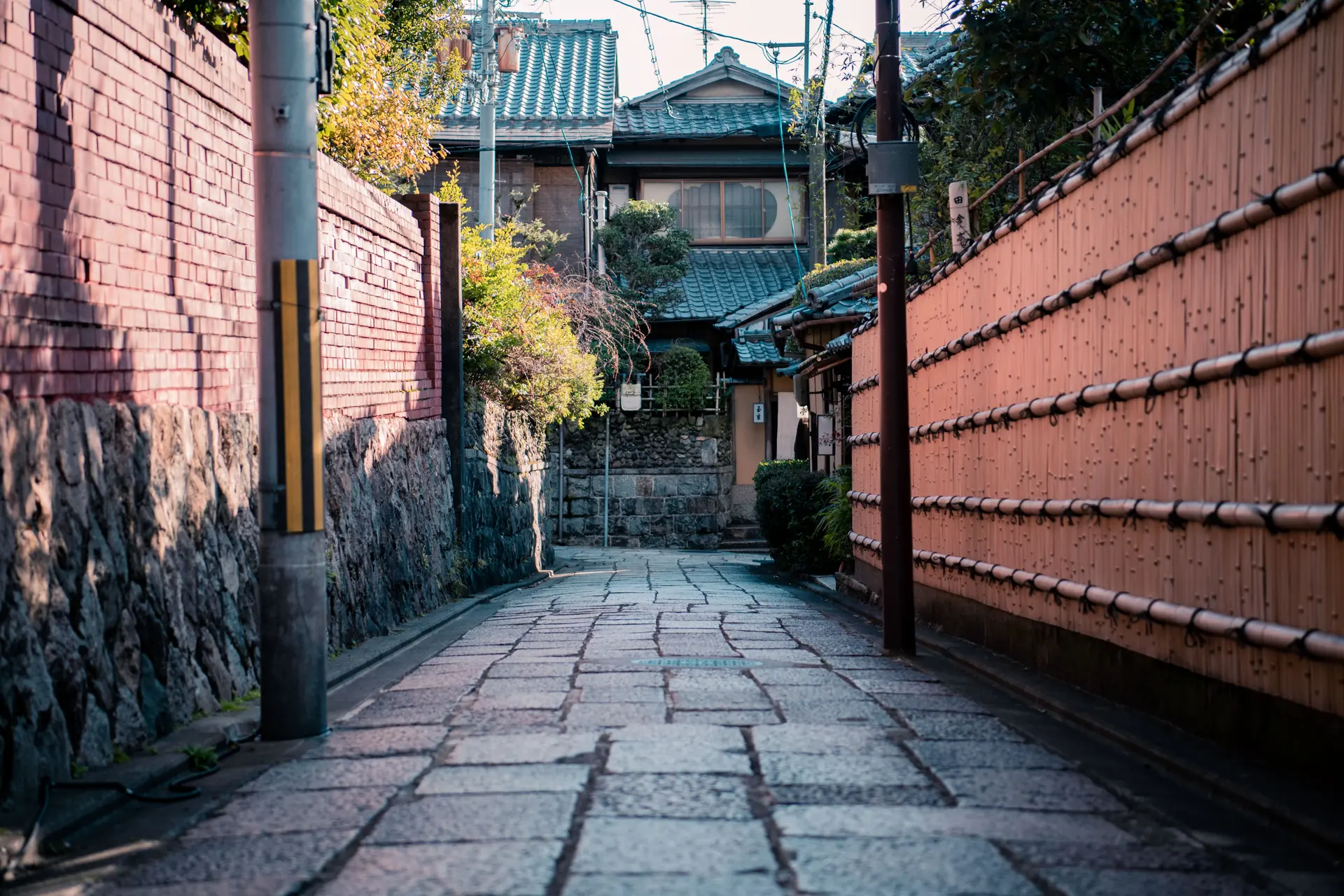 A stone-flagged Kyoto lane between a plaster wall and split-bamboo fencing, tiled machiya roofs ahead