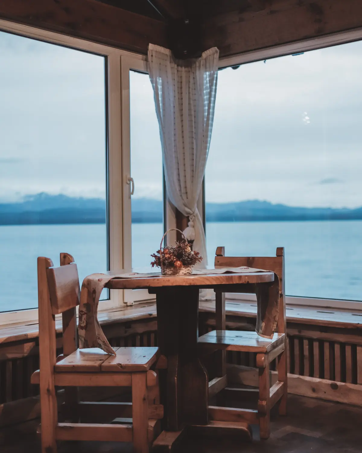 A timber-lined corner room with two carved chairs at three windows looking onto grey water and a snowy ridge