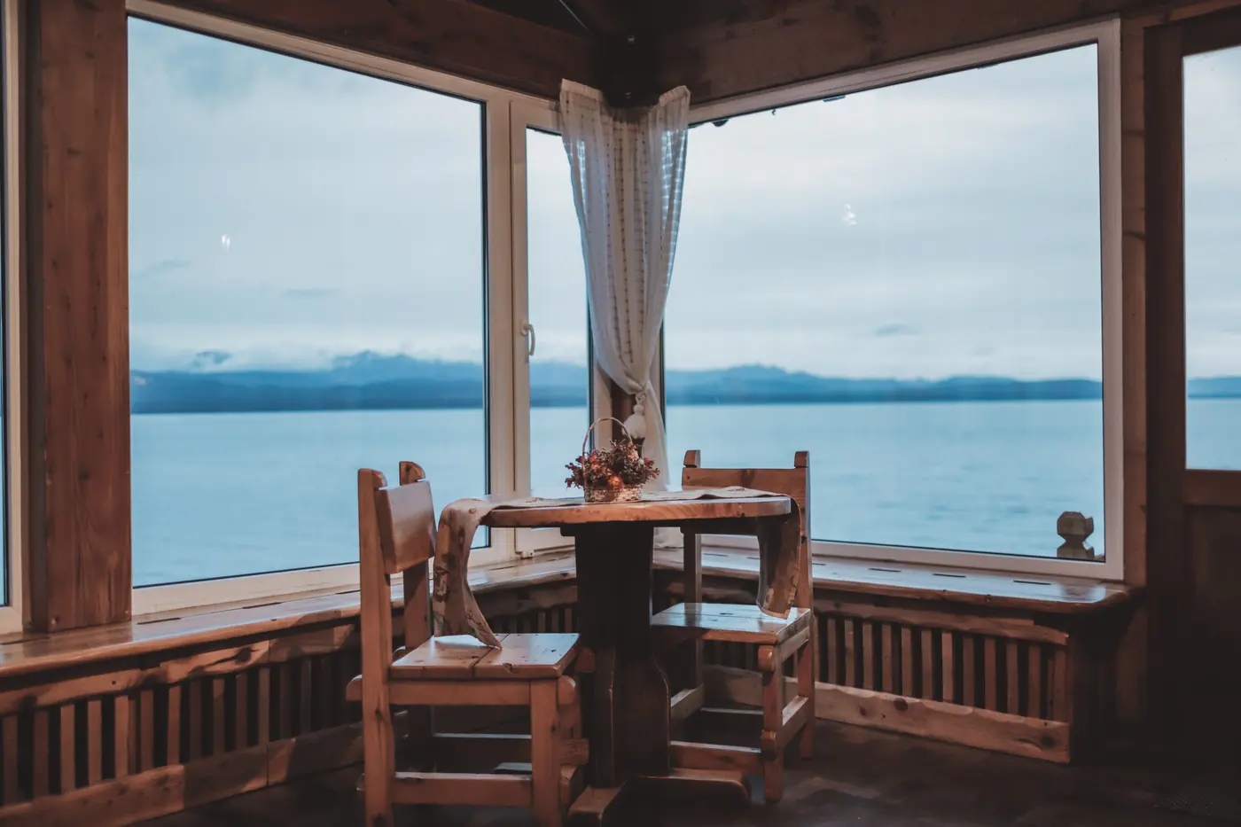 A timber-lined corner room with two carved chairs at three windows looking onto grey water and a snowy ridge