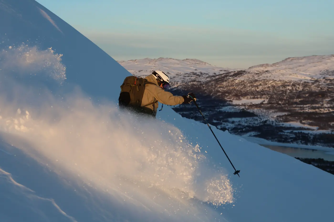 A skier in muted kit cutting a turn on an open snow slope above a fjord in low arctic sun