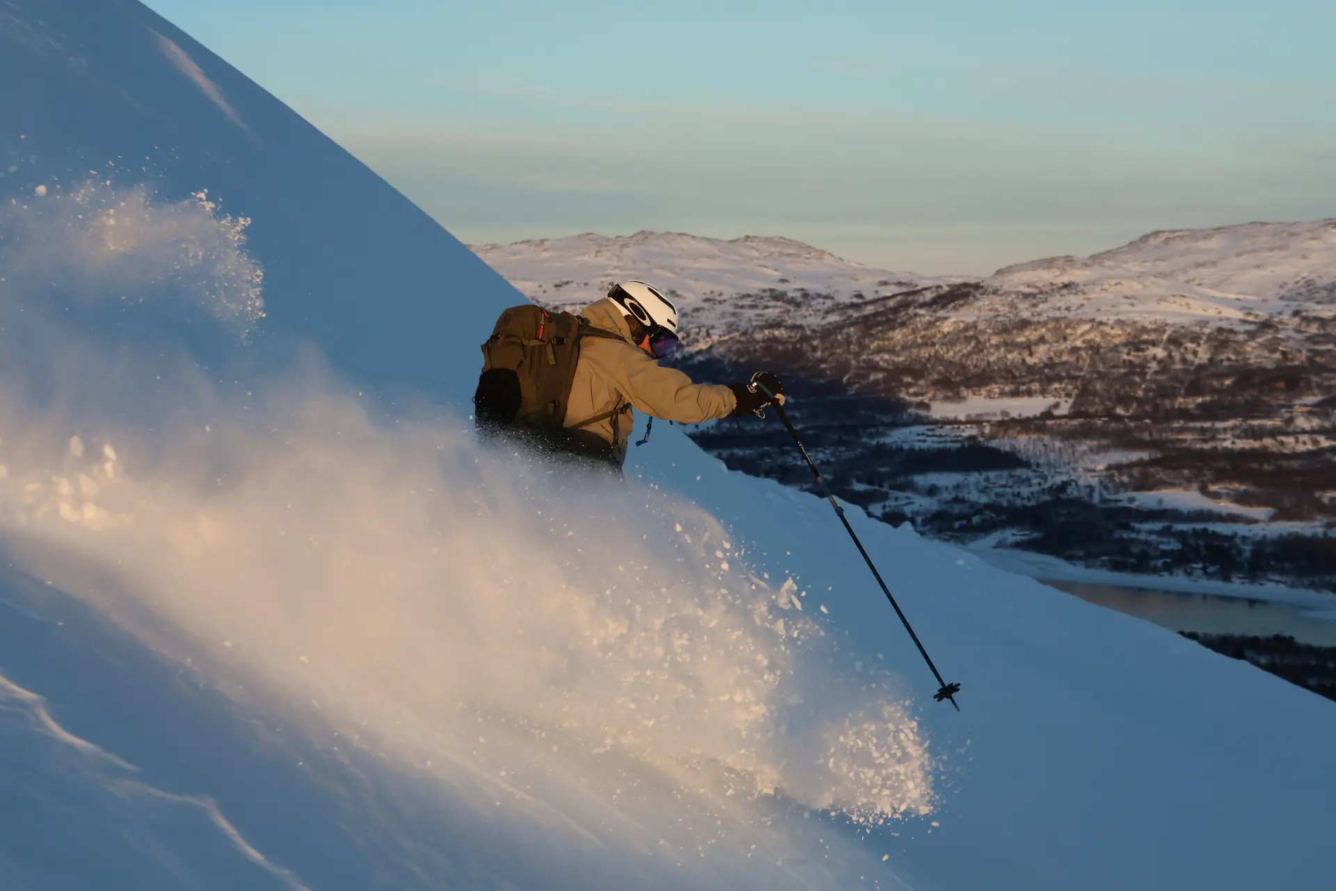 A skier in muted kit cutting a turn on an open snow slope above a fjord in low arctic sun