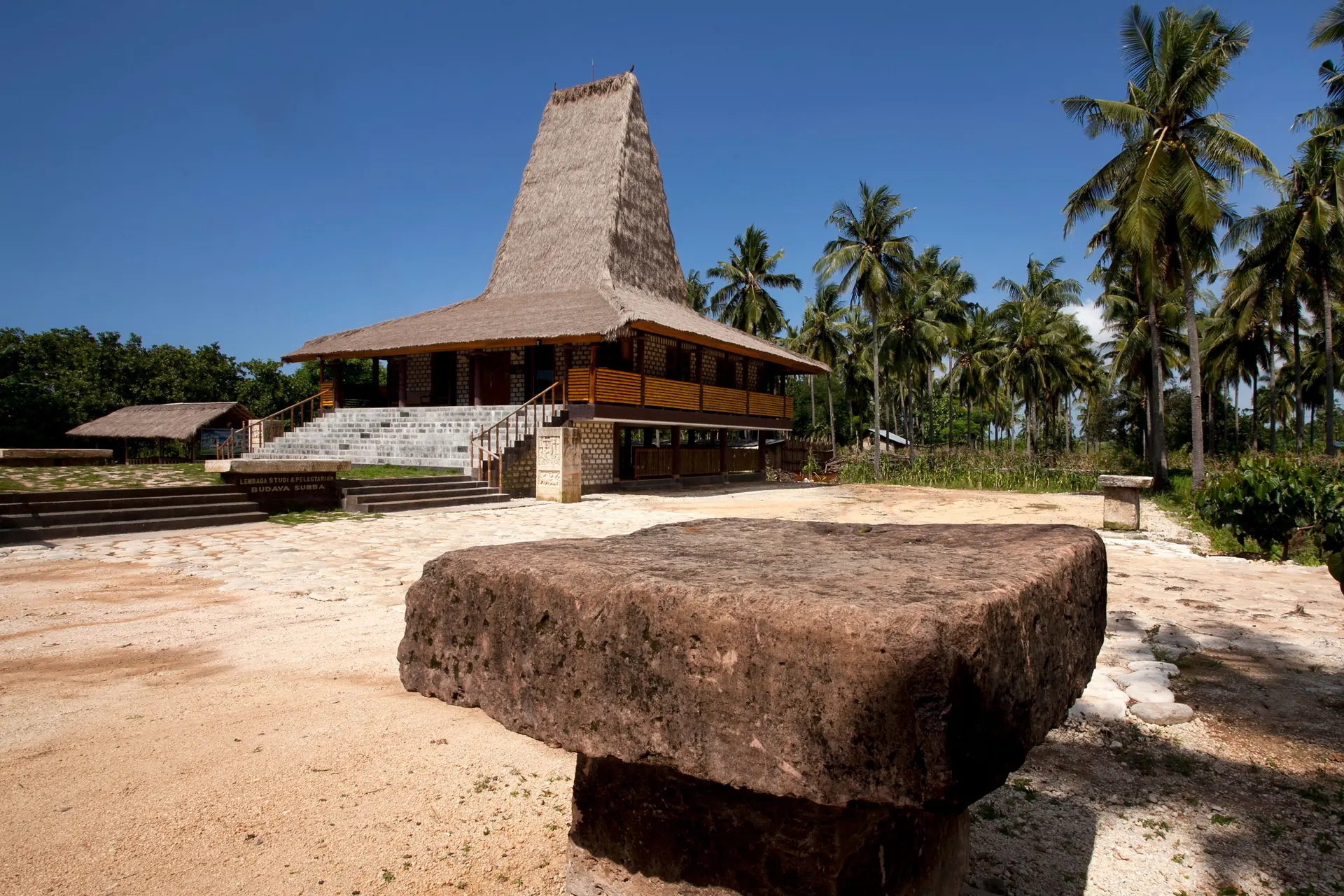 A high-peaked thatch clan house on timber posts behind a megalithic stone tomb on coral-sand paving