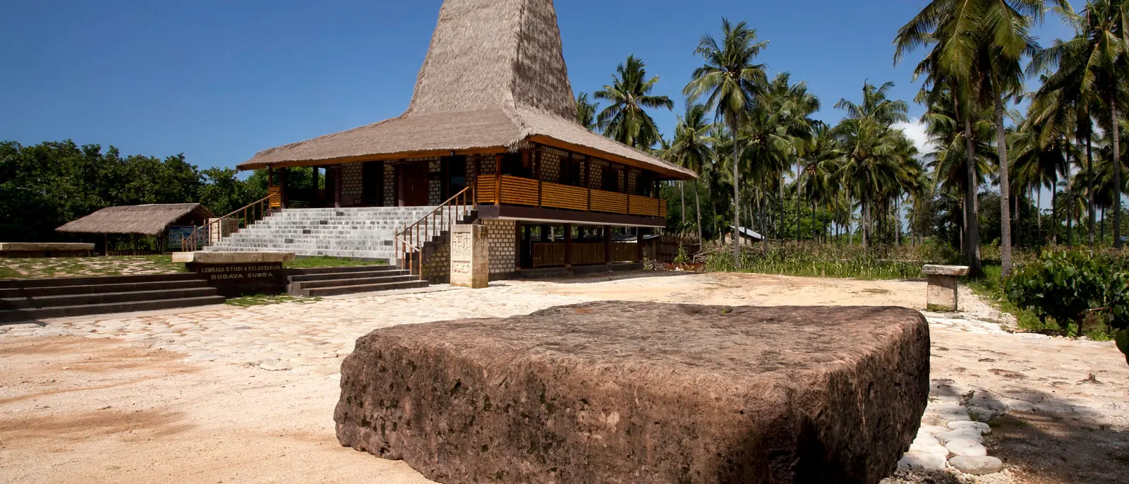A high-peaked thatch clan house on timber posts behind a megalithic stone tomb on coral-sand paving