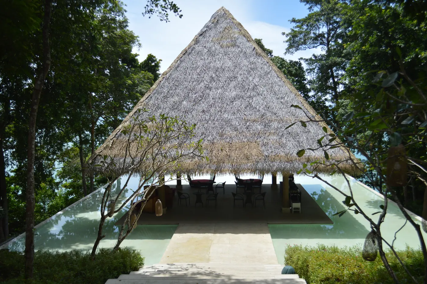 An open-sided pavilion under one steep grey thatch roof on a timber deck, sea visible beneath the eaves