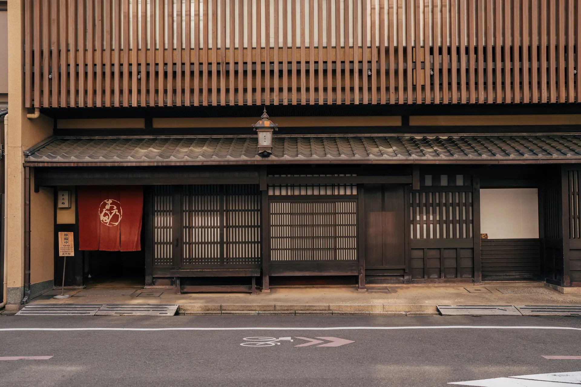 A machiya street front of fine cedar lattice with a tiled pent roof, paper lantern and indigo doorway curtain