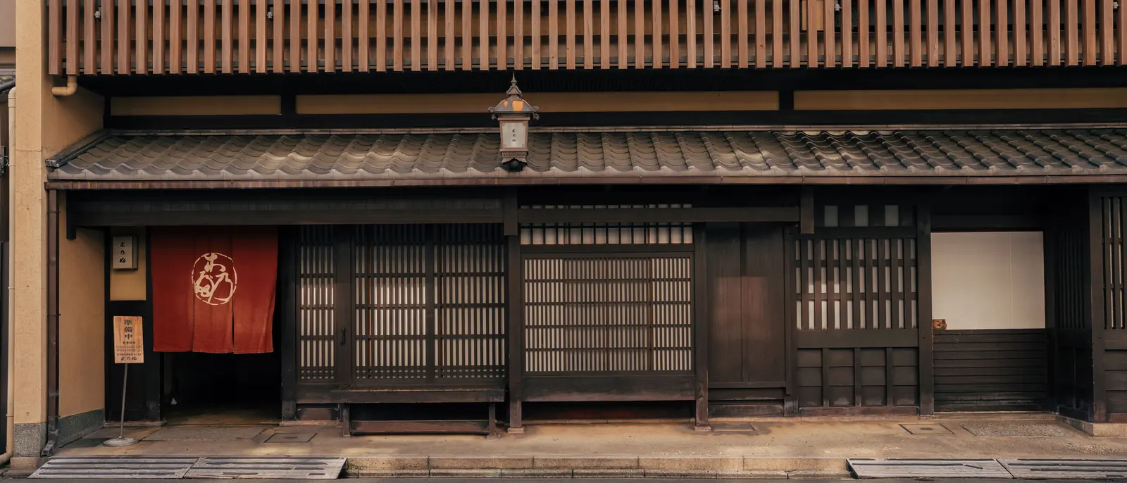 A machiya street front of fine cedar lattice with a tiled pent roof, paper lantern and indigo doorway curtain