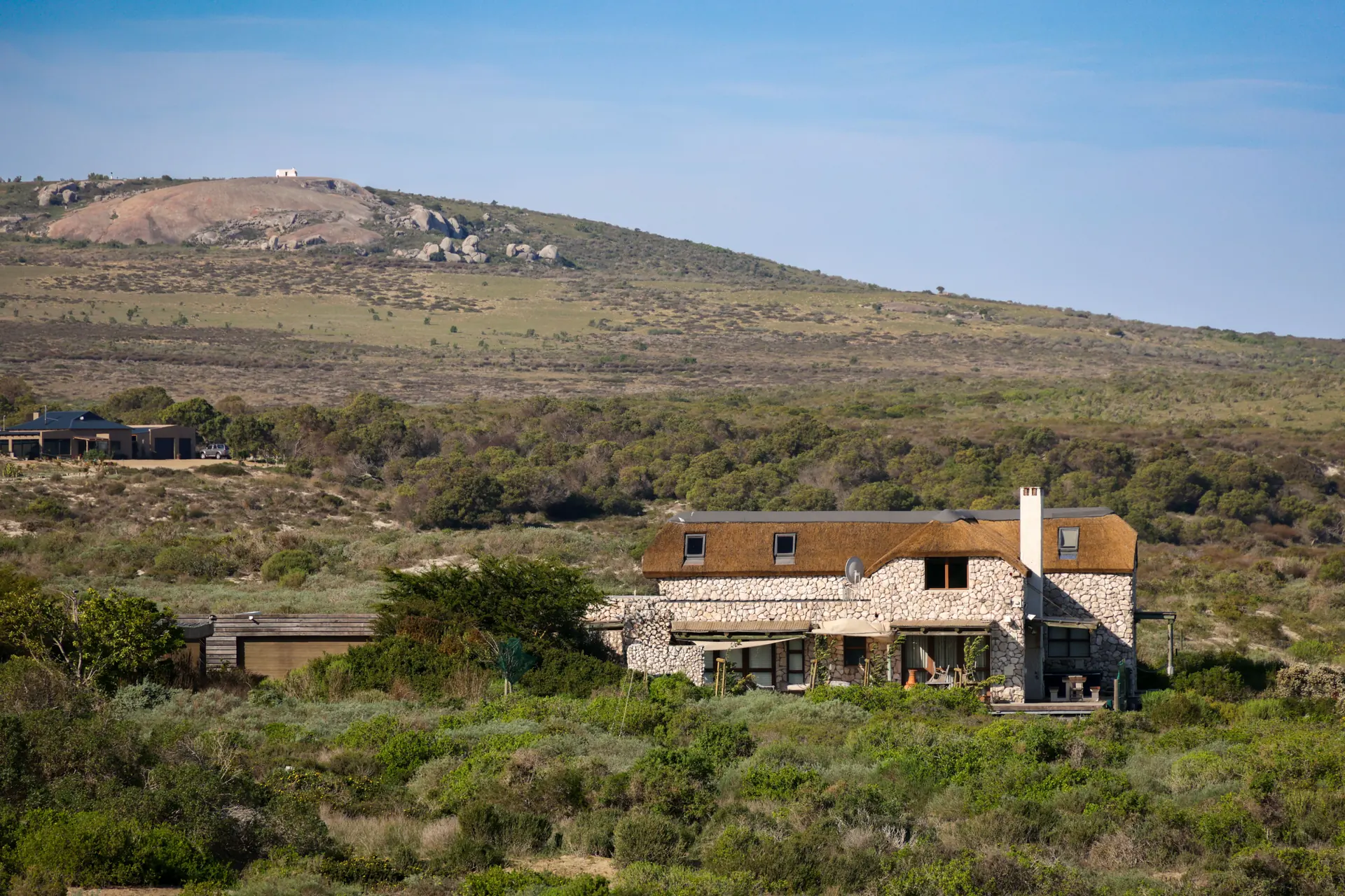 A low stone house with a steep shingled roof set into a scrub-covered hillside below a long rocky ridge