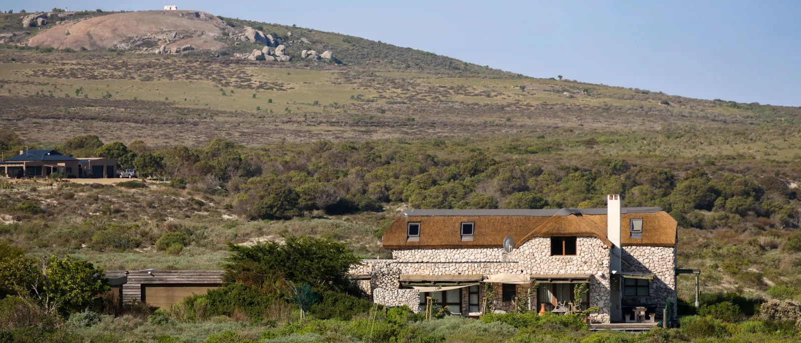 A low stone house with a steep shingled roof set into a scrub-covered hillside below a long rocky ridge