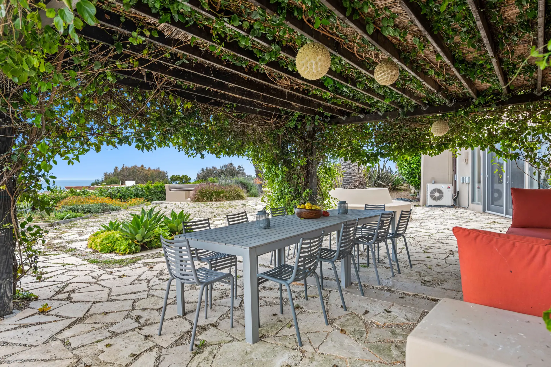 A vine-covered pergola over a stone terrace with one long table and eight chairs, the sea beyond