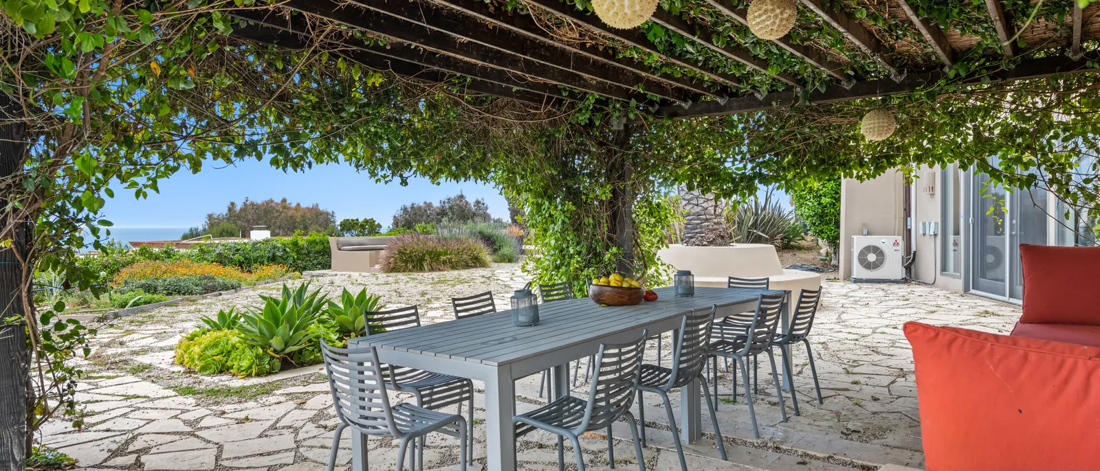 A vine-covered pergola over a stone terrace with one long table and eight chairs, the sea beyond