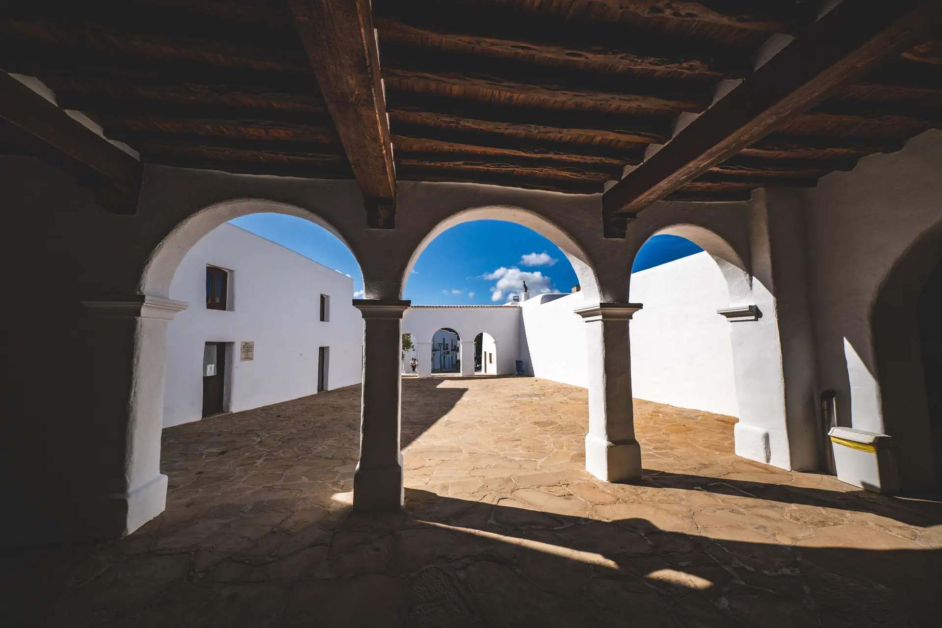 Whitewashed arches on heavy piers under a cane-and-beam ceiling, opening onto a sunlit flagstone courtyard