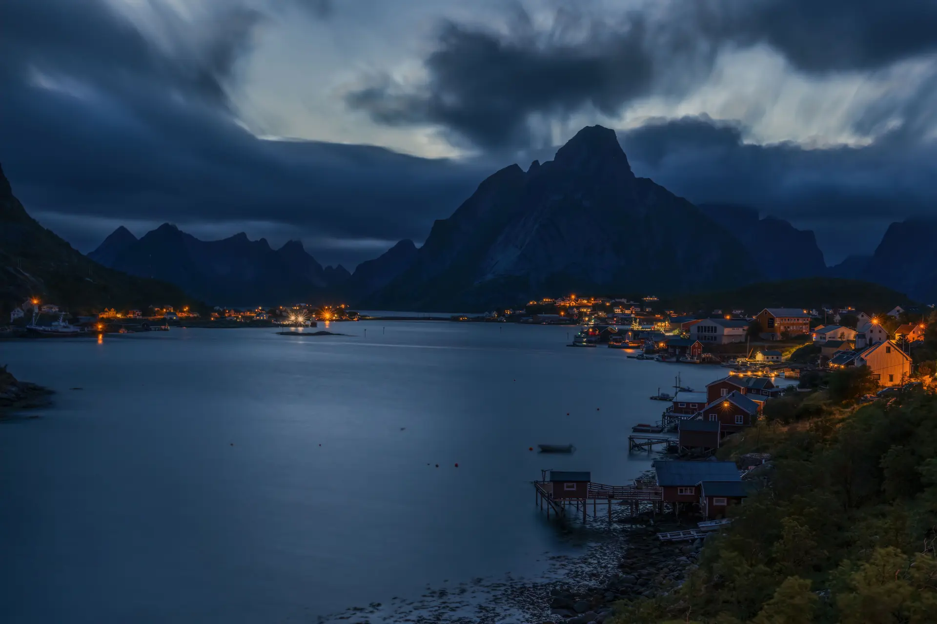 A serrated snowy granite ridge above a sound at blue hour, with lit cabins on stilts over the water